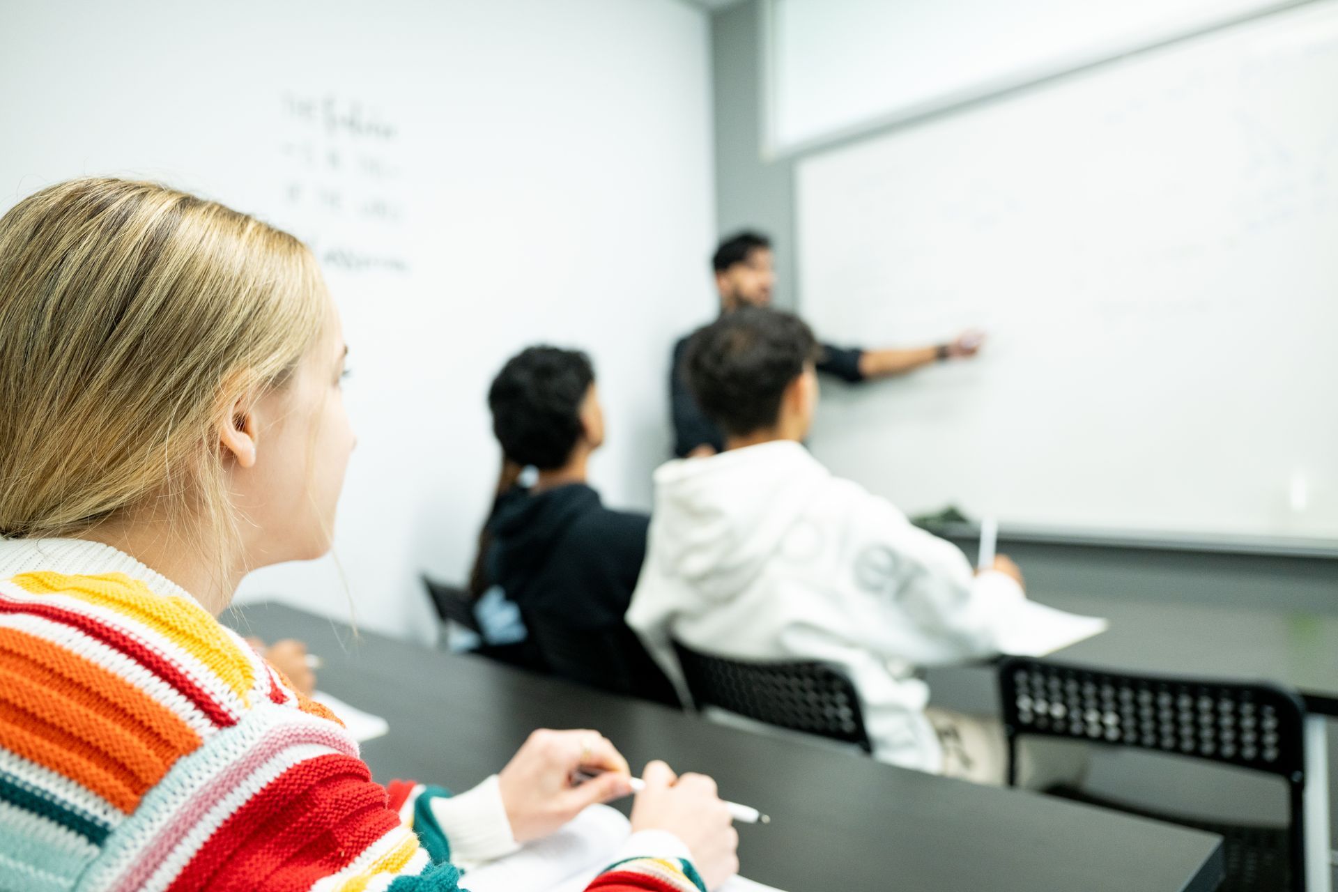 Students in a classroom listening to a teacher pointing at a whiteboard