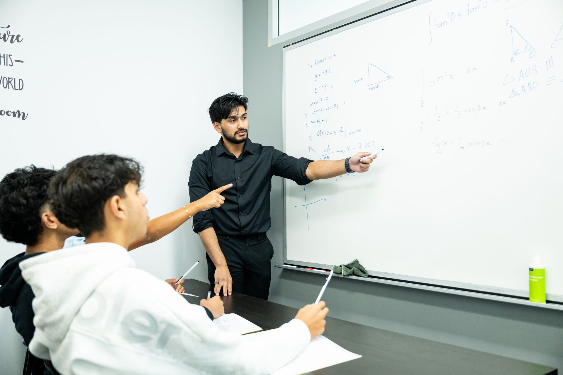 Teacher presenting at a whiteboard while two students listen in a classroom