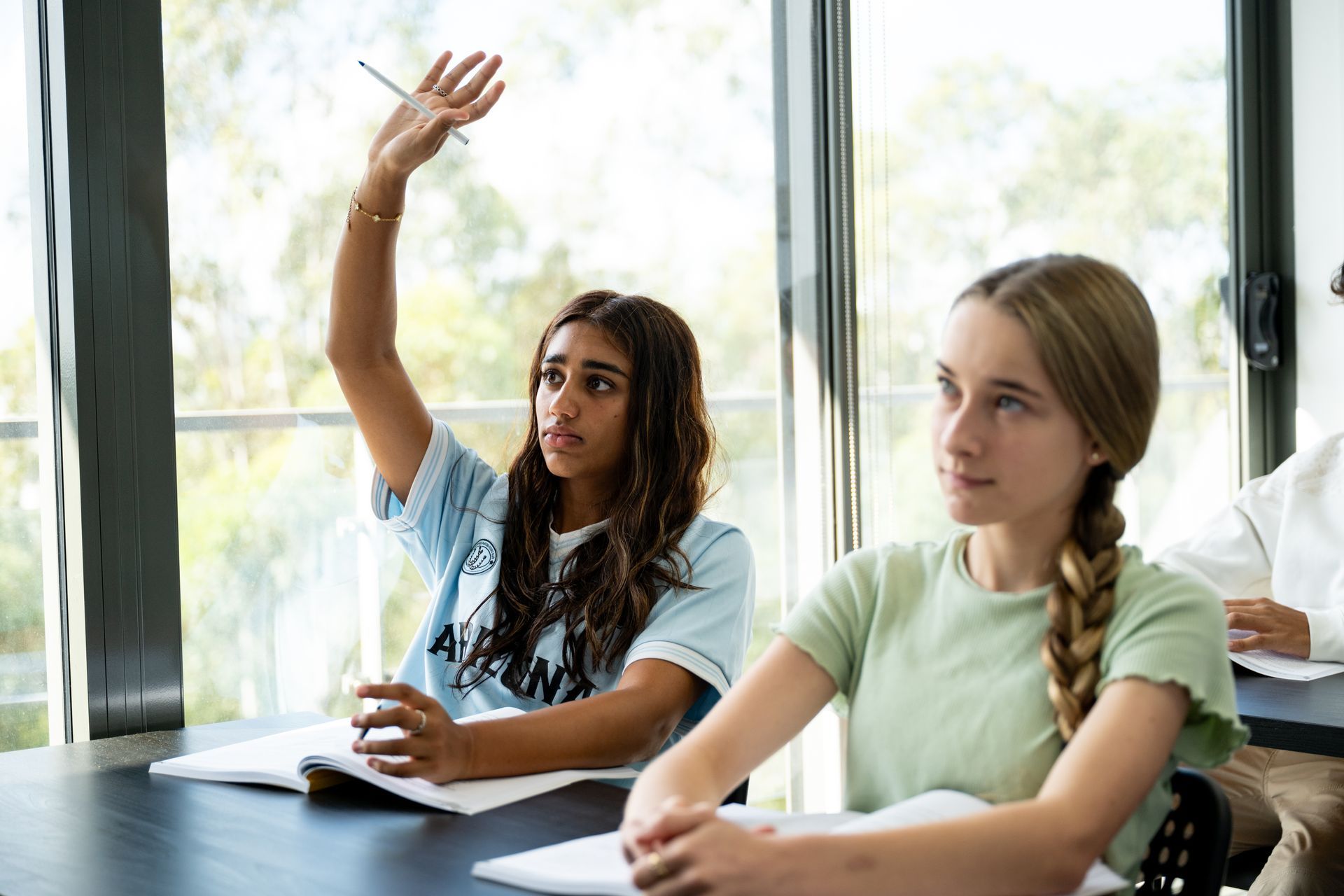 Students in a classroom, one raising a hand while others write at desks.