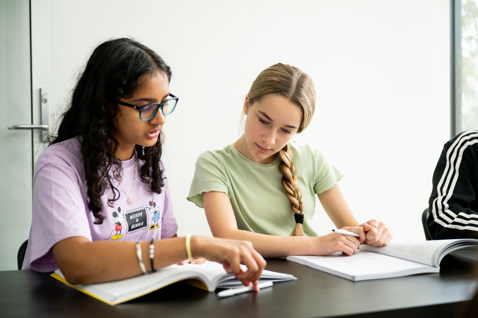 Two students studying together at a table, writing in notebooks with an open book nearby.
