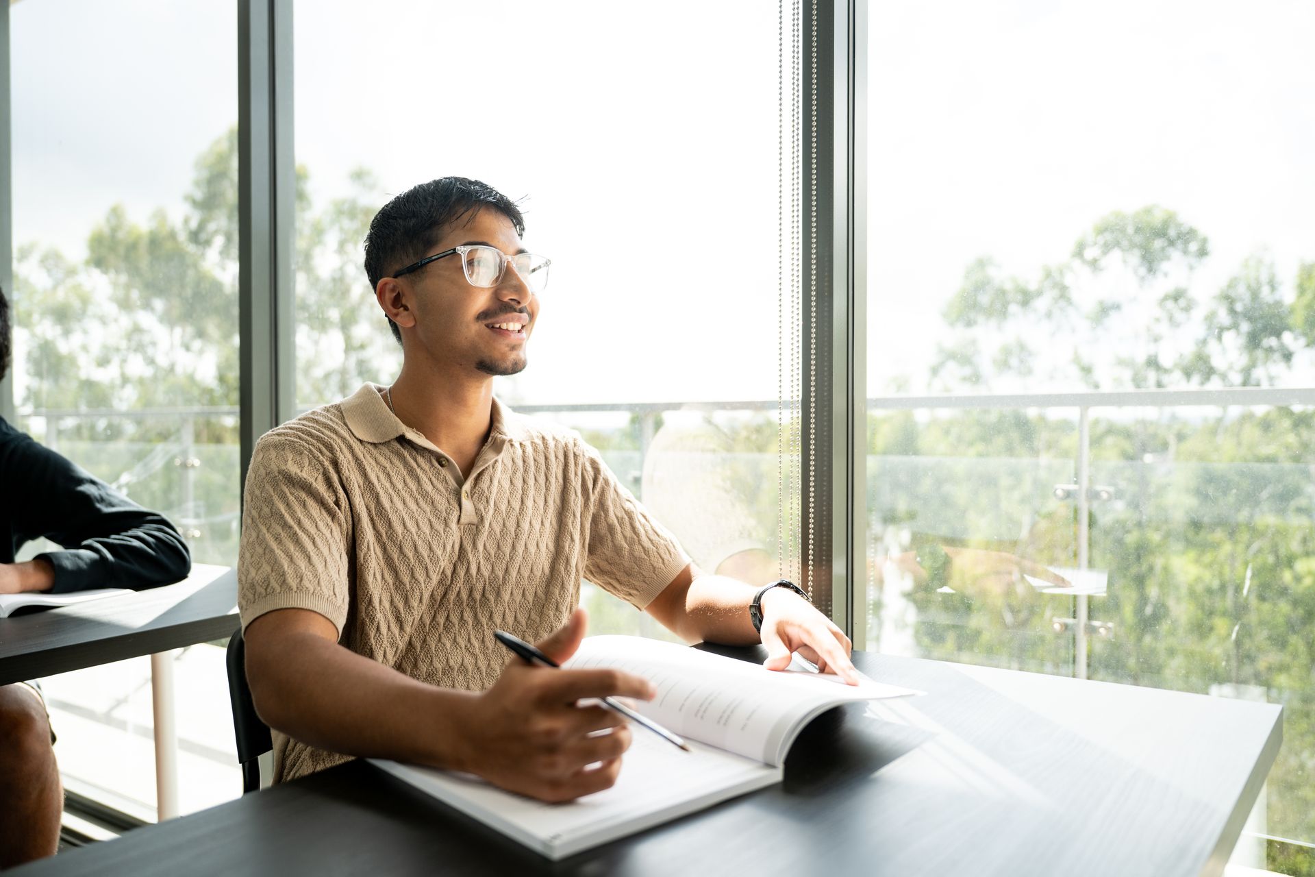 Man reading an open notebook at a table by large windows in a bright room