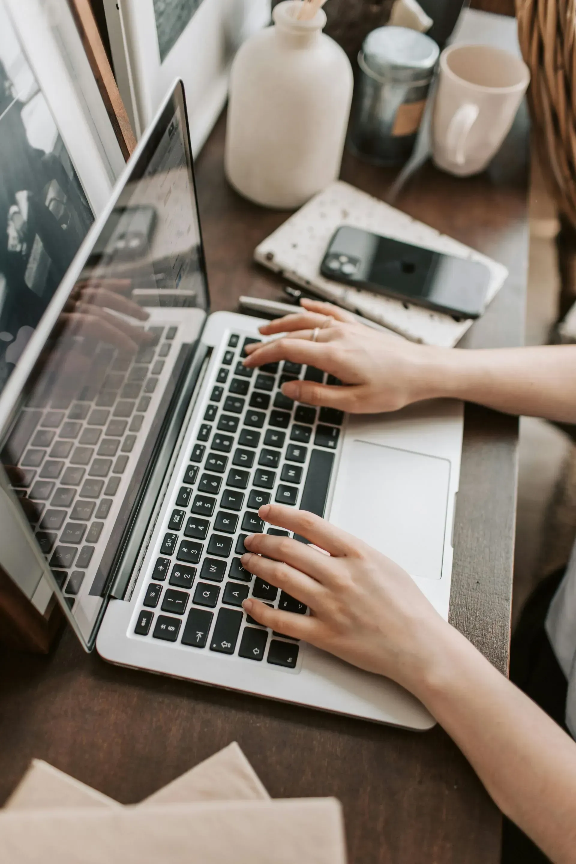 Hands typing on a laptop at a wooden desk with a phone, notebook, and vase nearby