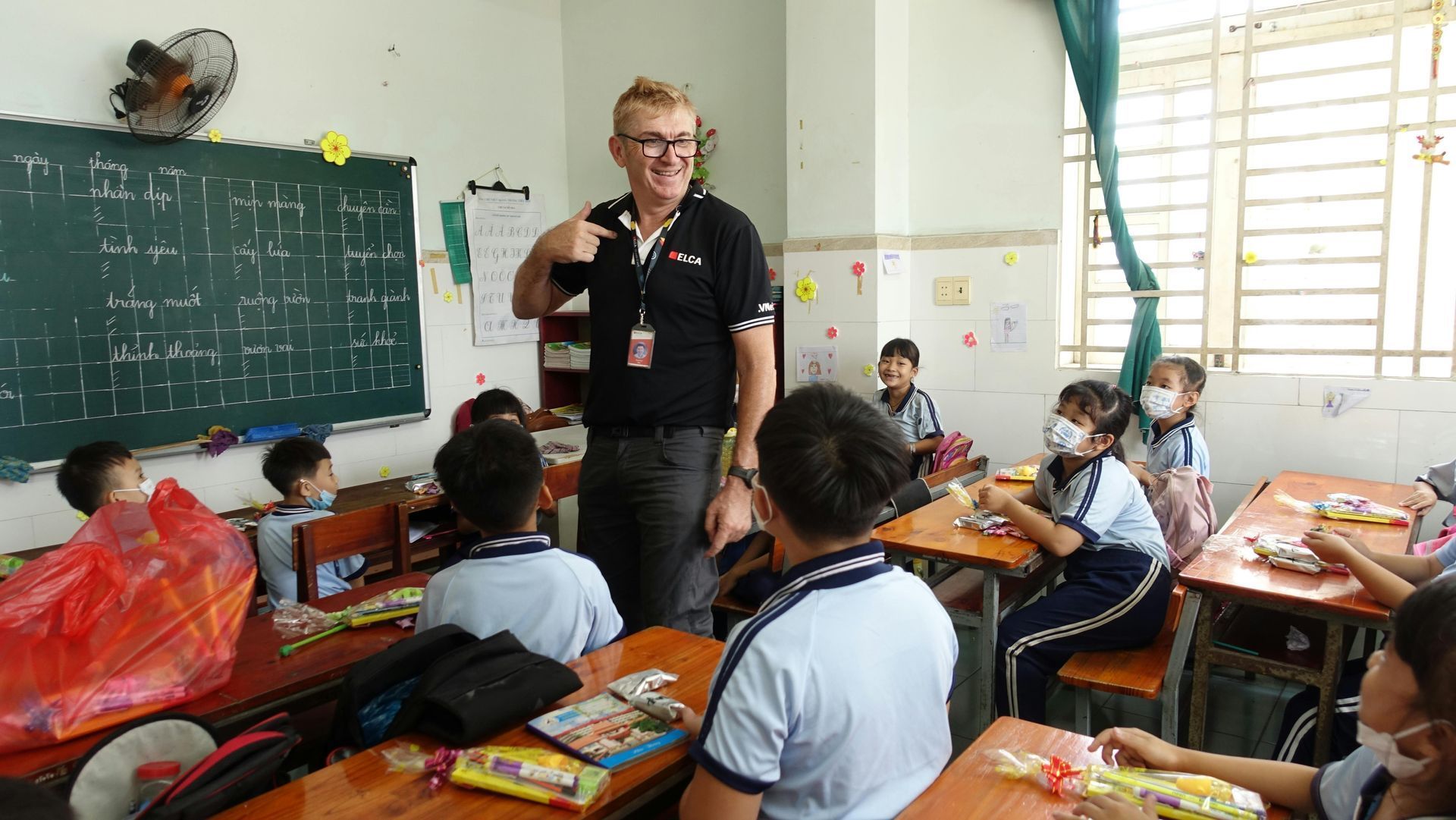 Teacher speaking to students in a classroom with chalkboard, desks, and windows