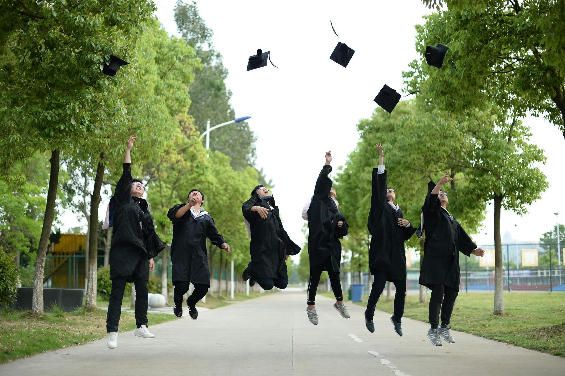 Group of students in black outfits jumping and tossing graduation caps on a tree-lined walkway.