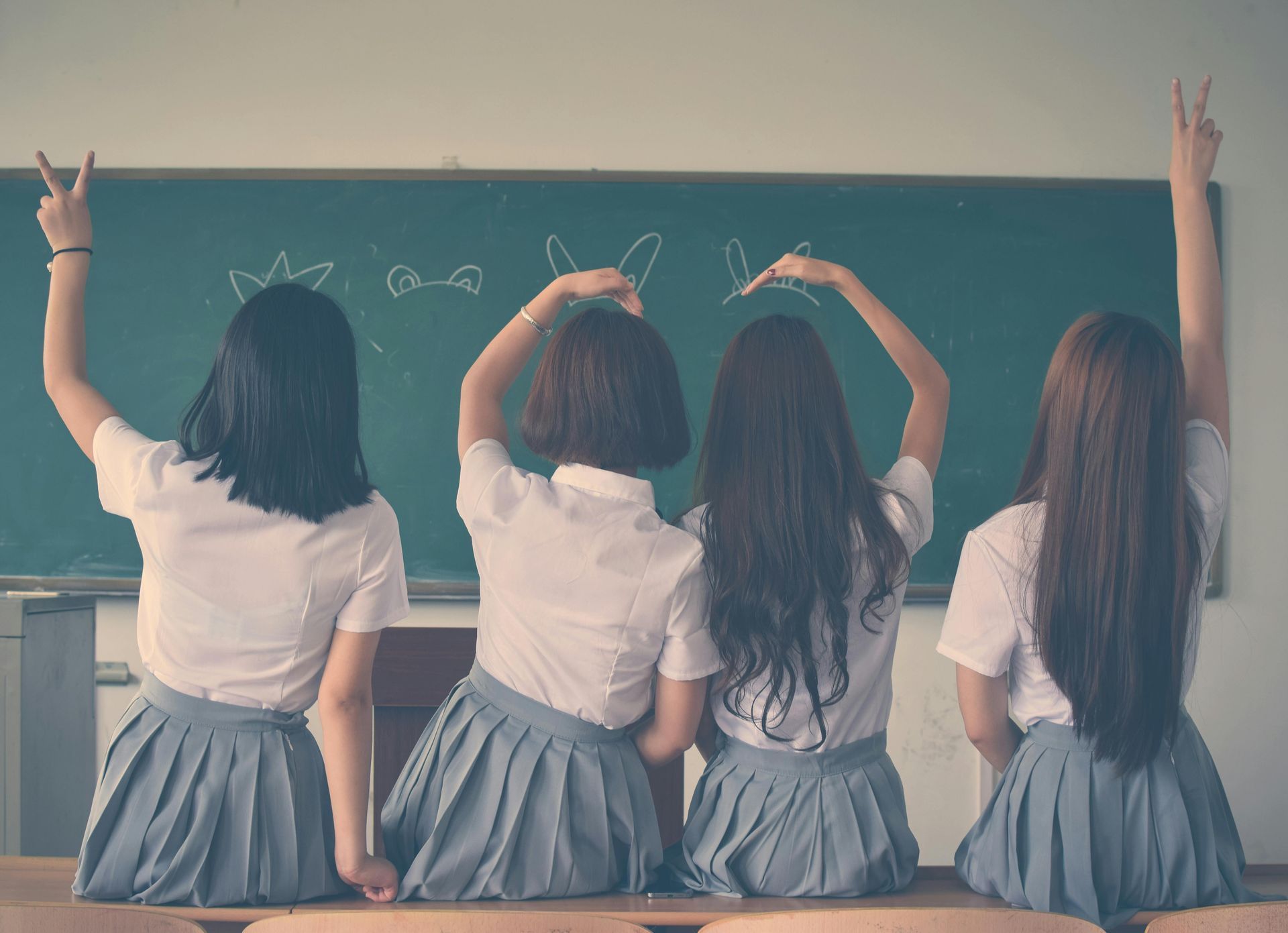 Four students in white shirts and gray skirts sit facing a chalkboard