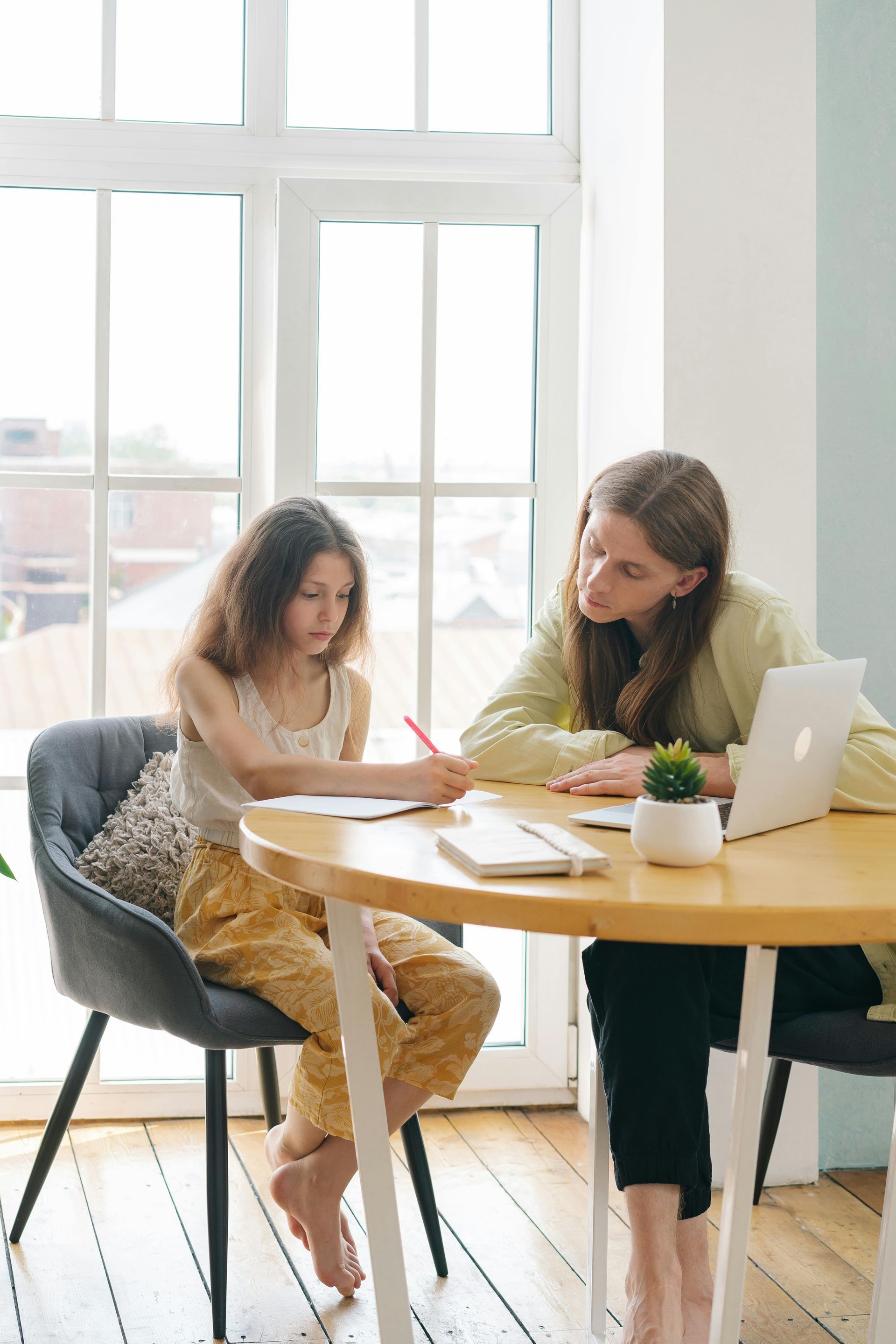 Two women review papers at a round table by a bright window, with a laptop and notebook nearby.