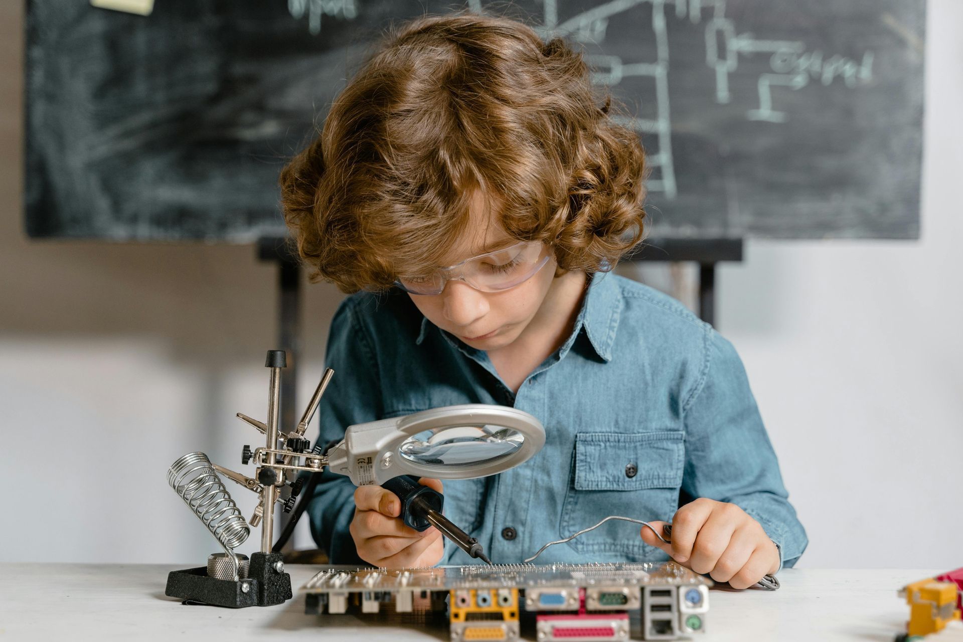 Child using a magnifying glass to inspect electronics on a workbench in a classroom or lab