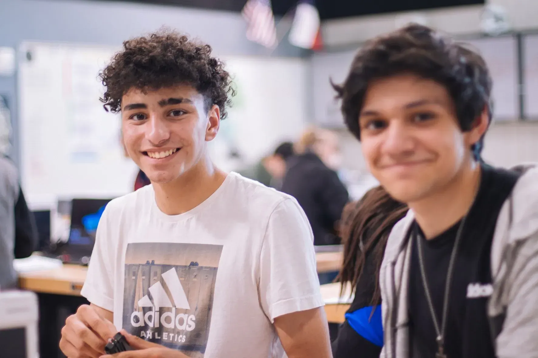 Two smiling students in a classroom, one in a white Adidas shirt and one in a black hoodie.
