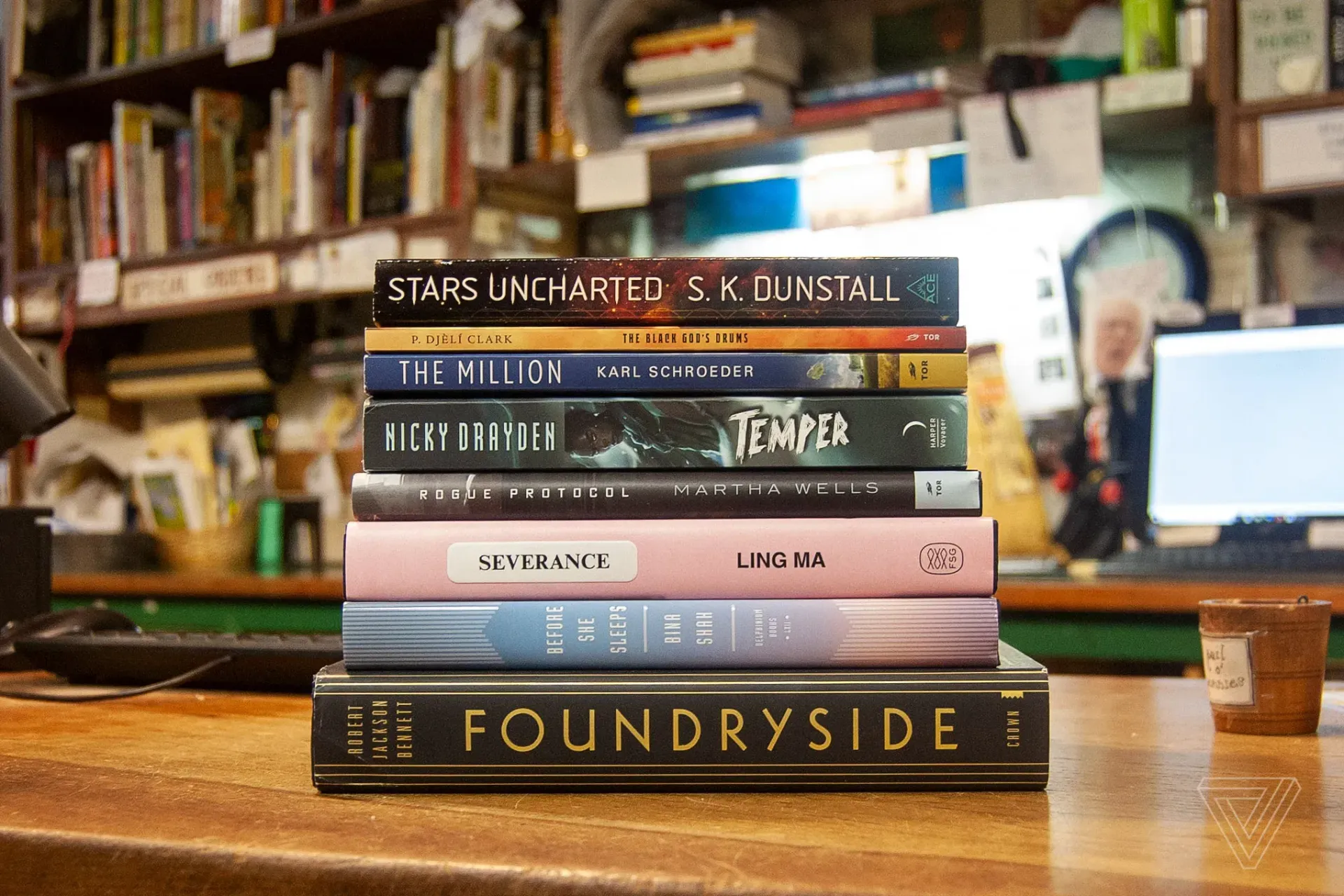 A stack of seven books rests on a wooden counter in front of a blurred bookshelf and computer screen.