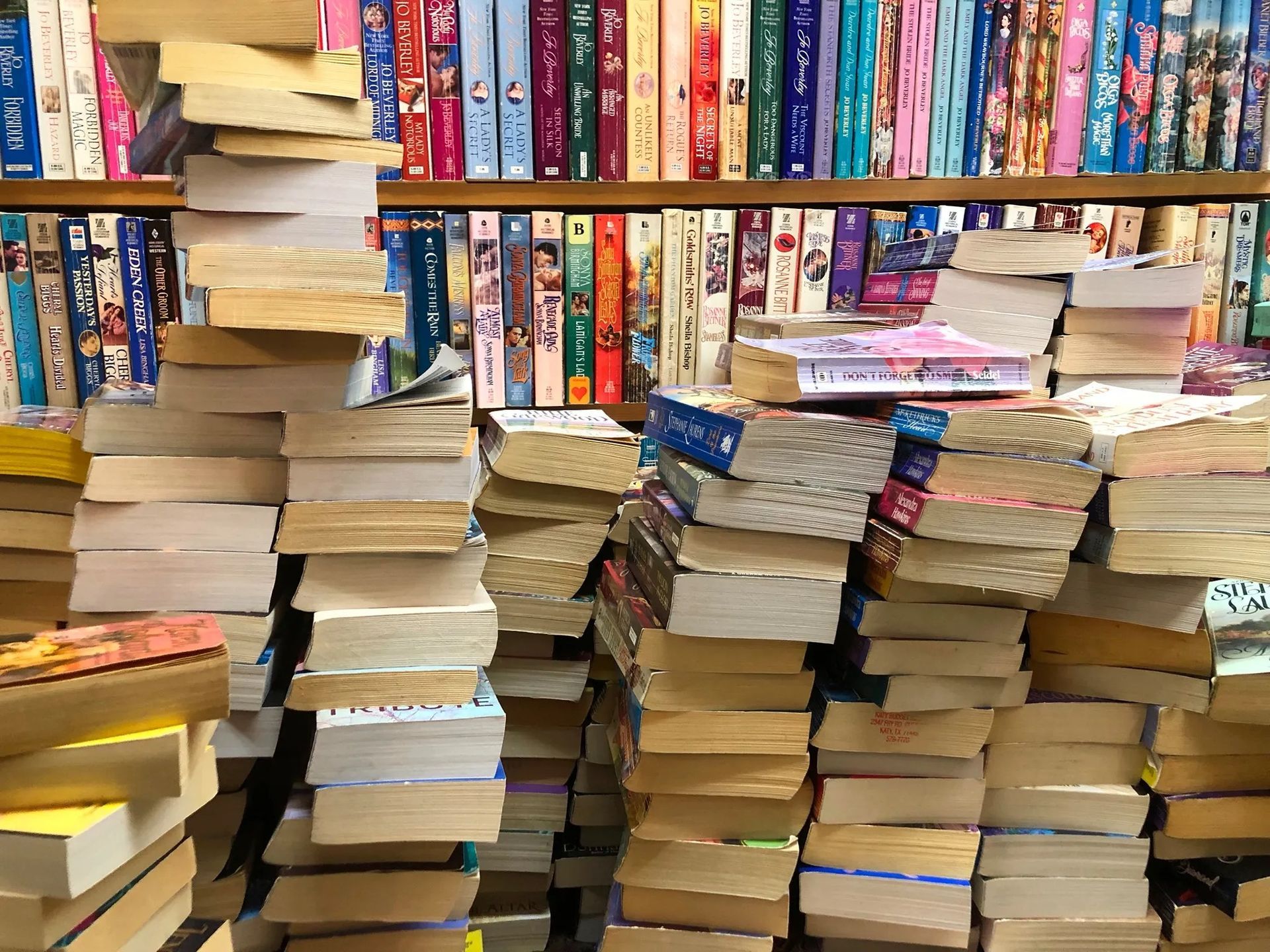 Stacks of books piled in front of colorful bookshelves in a bookstore or library.