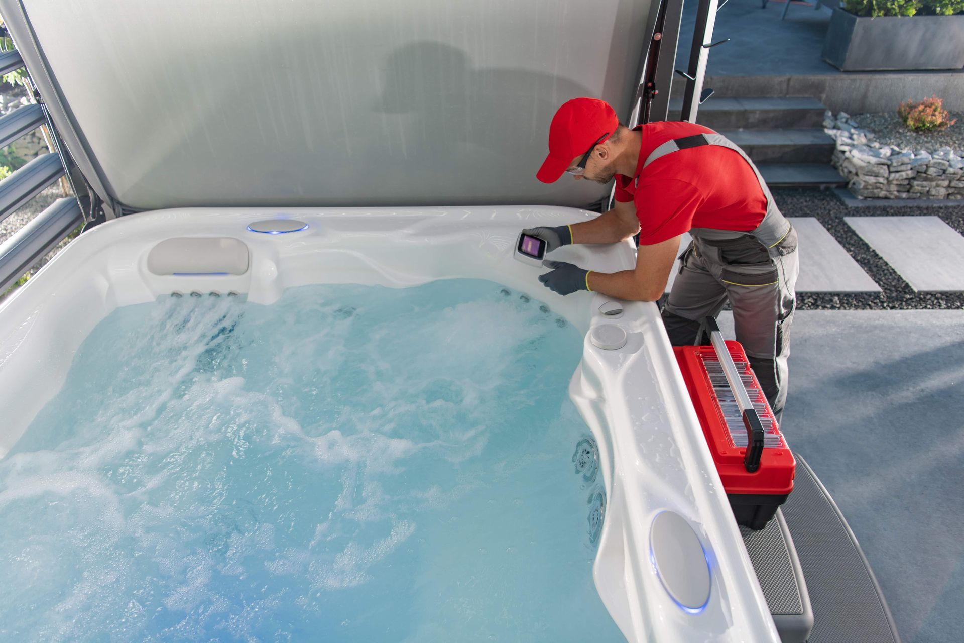Man in red shirt and hat repairs hot tub with tool kit; outdoors.