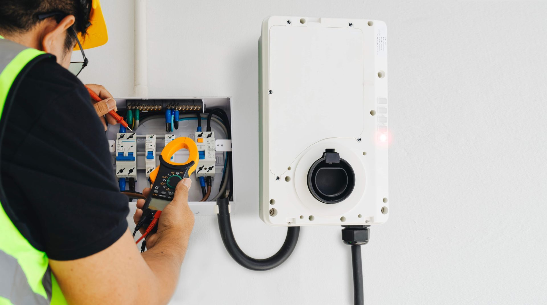Electrician in yellow vest uses a clamp meter to check wiring on a white wall with an EV charger.
