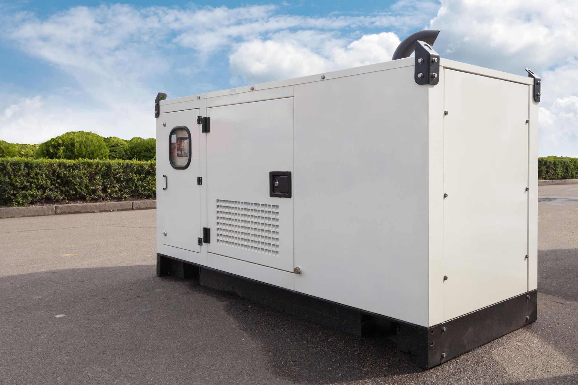 White generator in an outdoor setting; blue sky, hedges, and paved ground are visible.