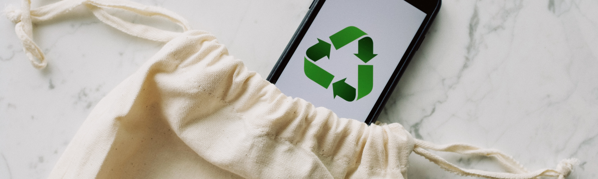 A smartphone displaying a green recycling symbol sits inside a light-colored fabric drawstring bag on a marble surface.