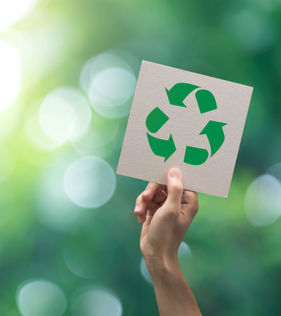 A hand holds up a square piece of paper with a green recycling symbol against a soft-focus green nature background.