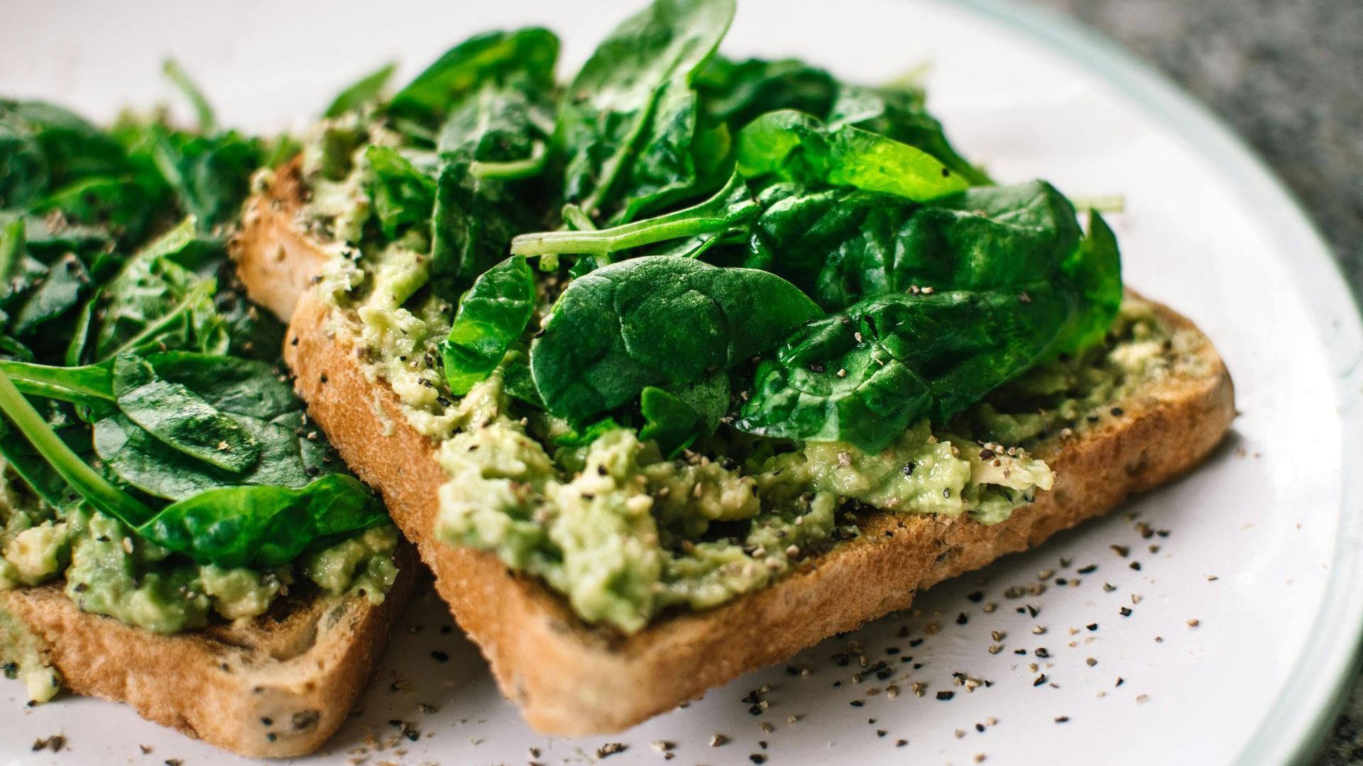 Two slices of toast with avocado and spinach on a white plate.
