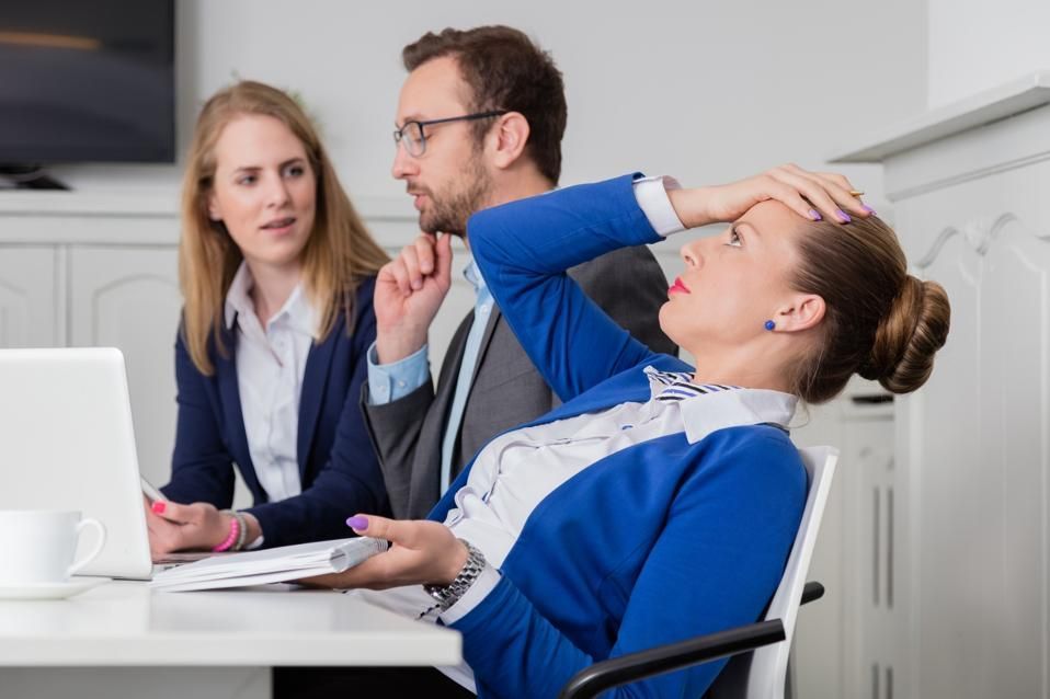 Three coworkers in a meeting; one woman leans back rubbing her forehead, looking stressed.