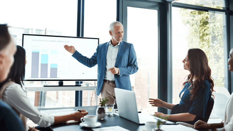 Business meeting with presenter showing charts on a screen in a bright office conference room