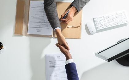 Two people in business attire shaking hands across a white desk with paperwork and a keyboard.