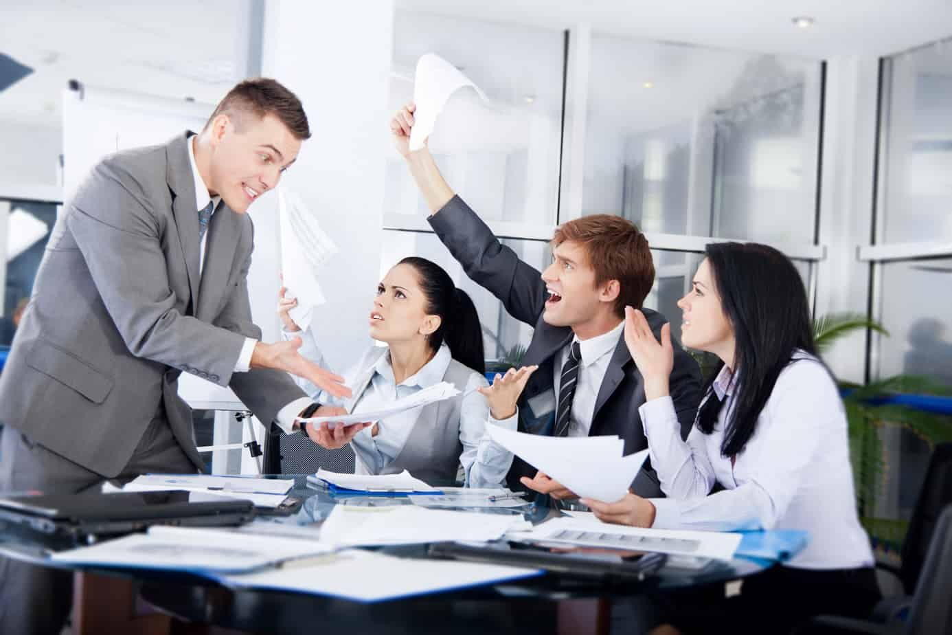 Business meeting in a bright office, with colleagues discussing documents and one man standing and gesturing.