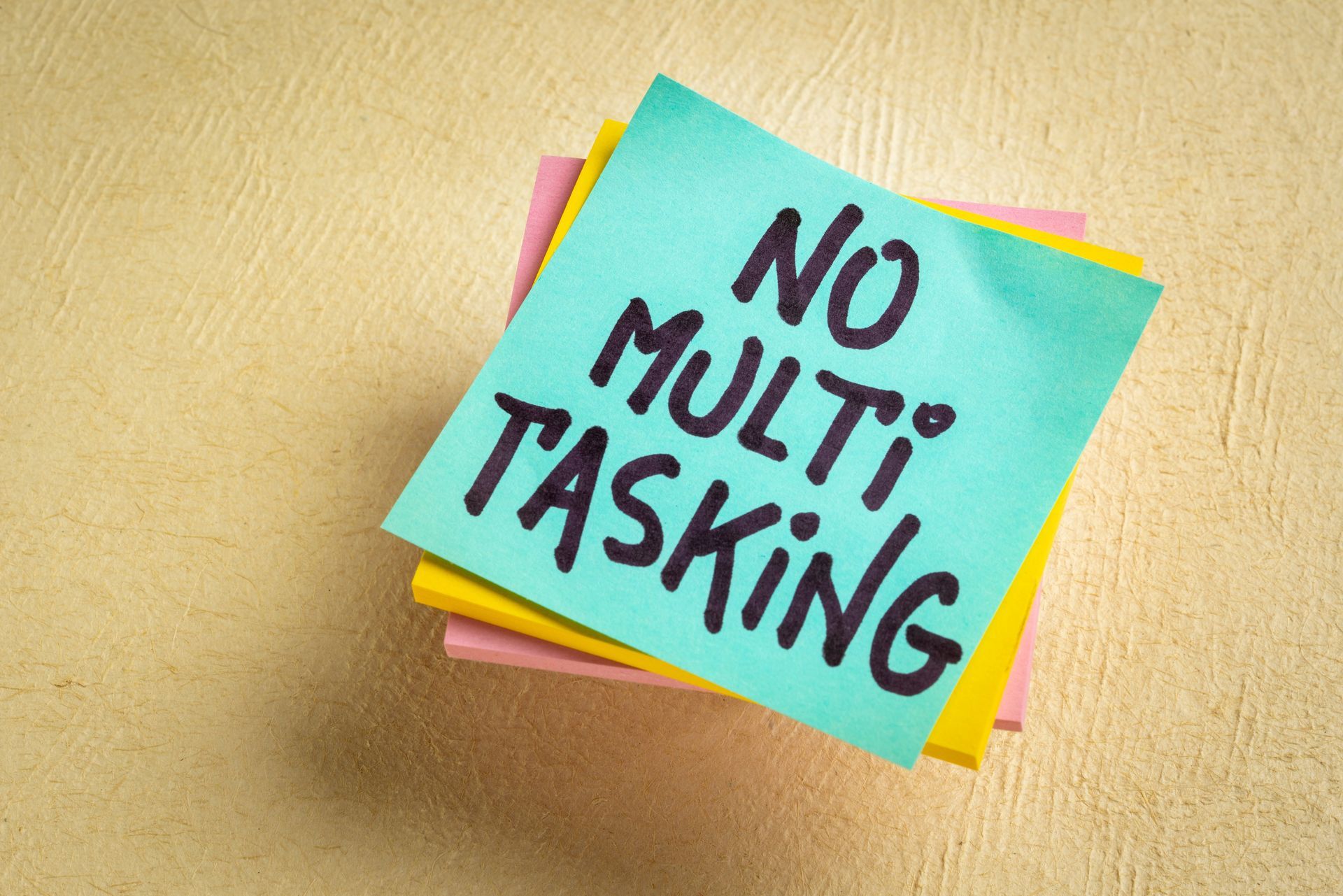 Colorful sticky notes with a turquoise note reading “NO MULTI-TASKING” on a tan surface