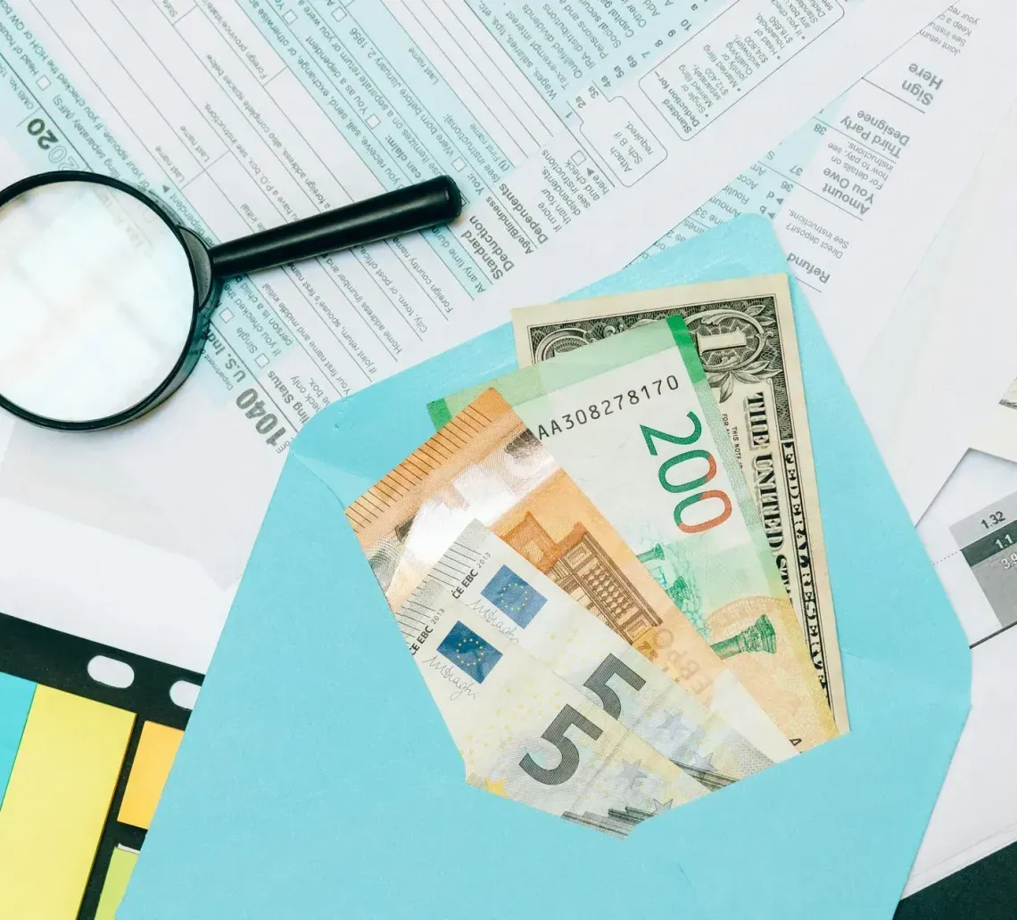 A light blue envelope holding Euro and Dollar bills, with a magnifying glass resting on financial tax forms.