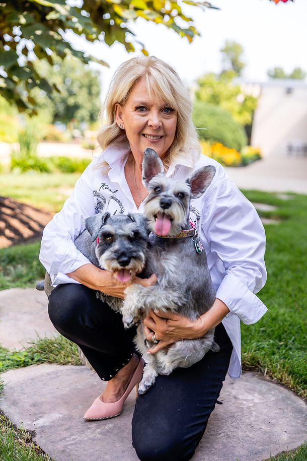 Woman kneeling on a garden path, holding two small dogs in a sunny yard.