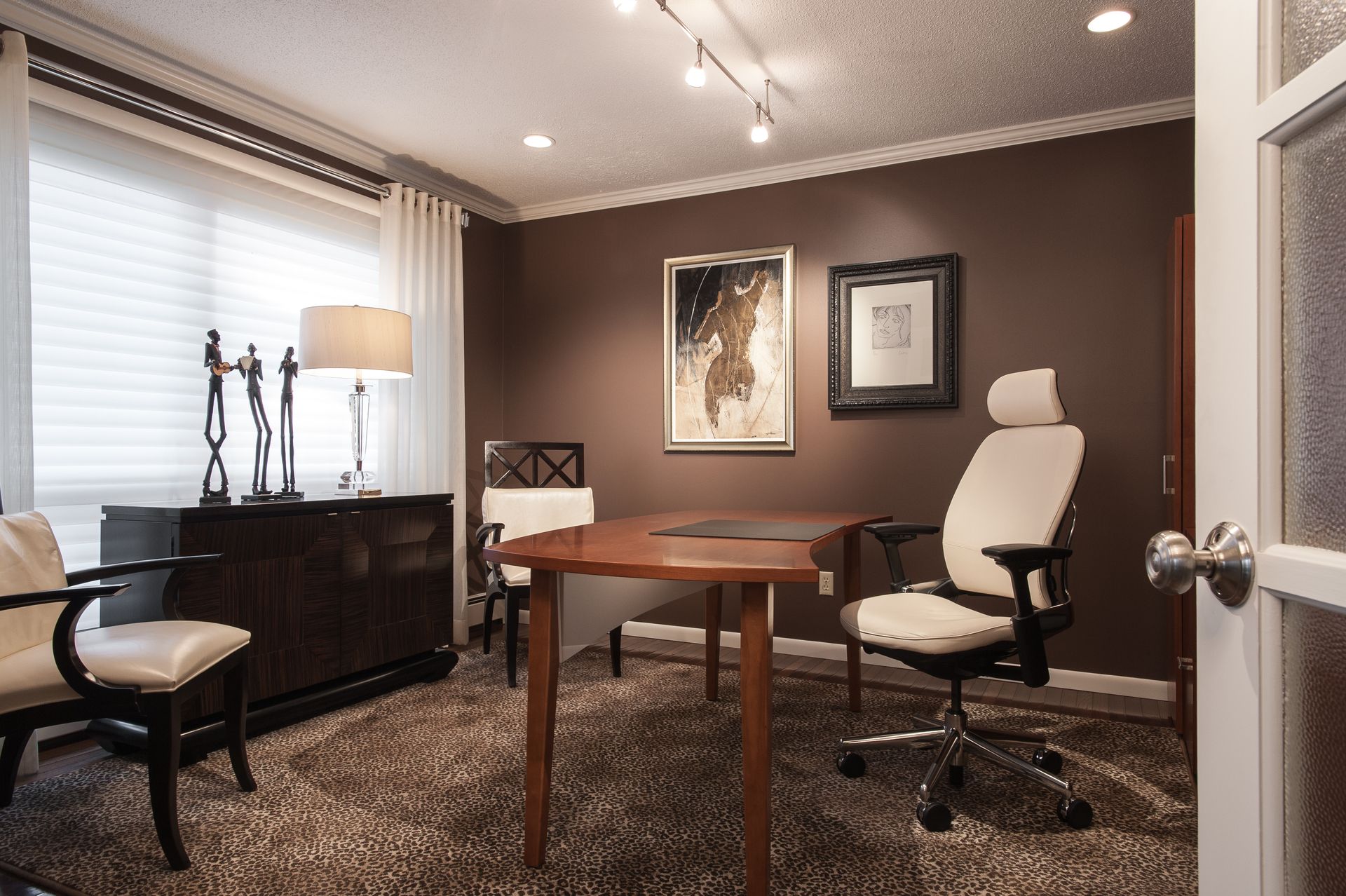 Modern home office with a wooden desk, white chair, dark cabinet, and wall art on brown walls.