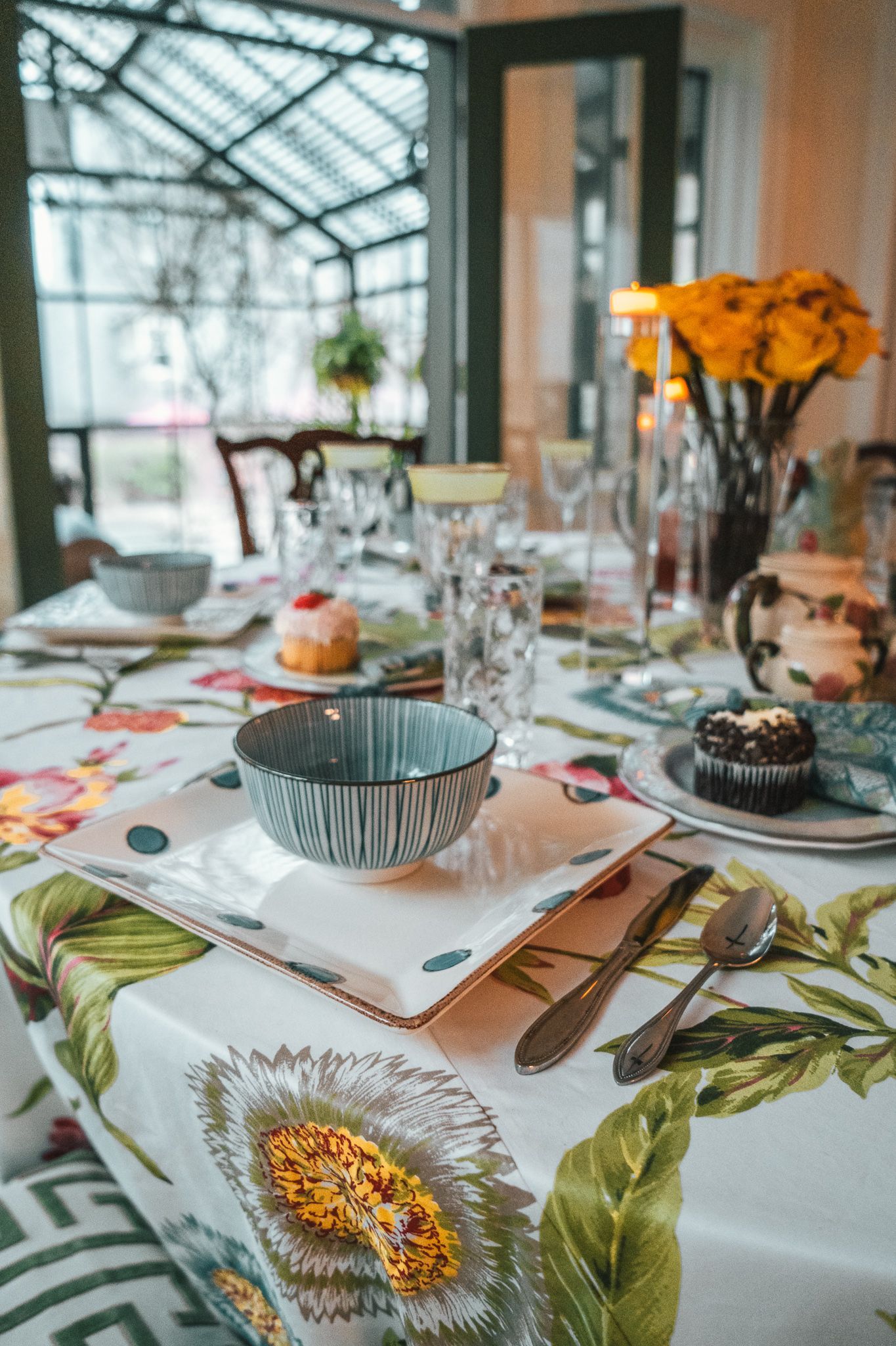 Decorated dining table with floral tablecloth, dishes, desserts, and orange flowers in a bright sunroom