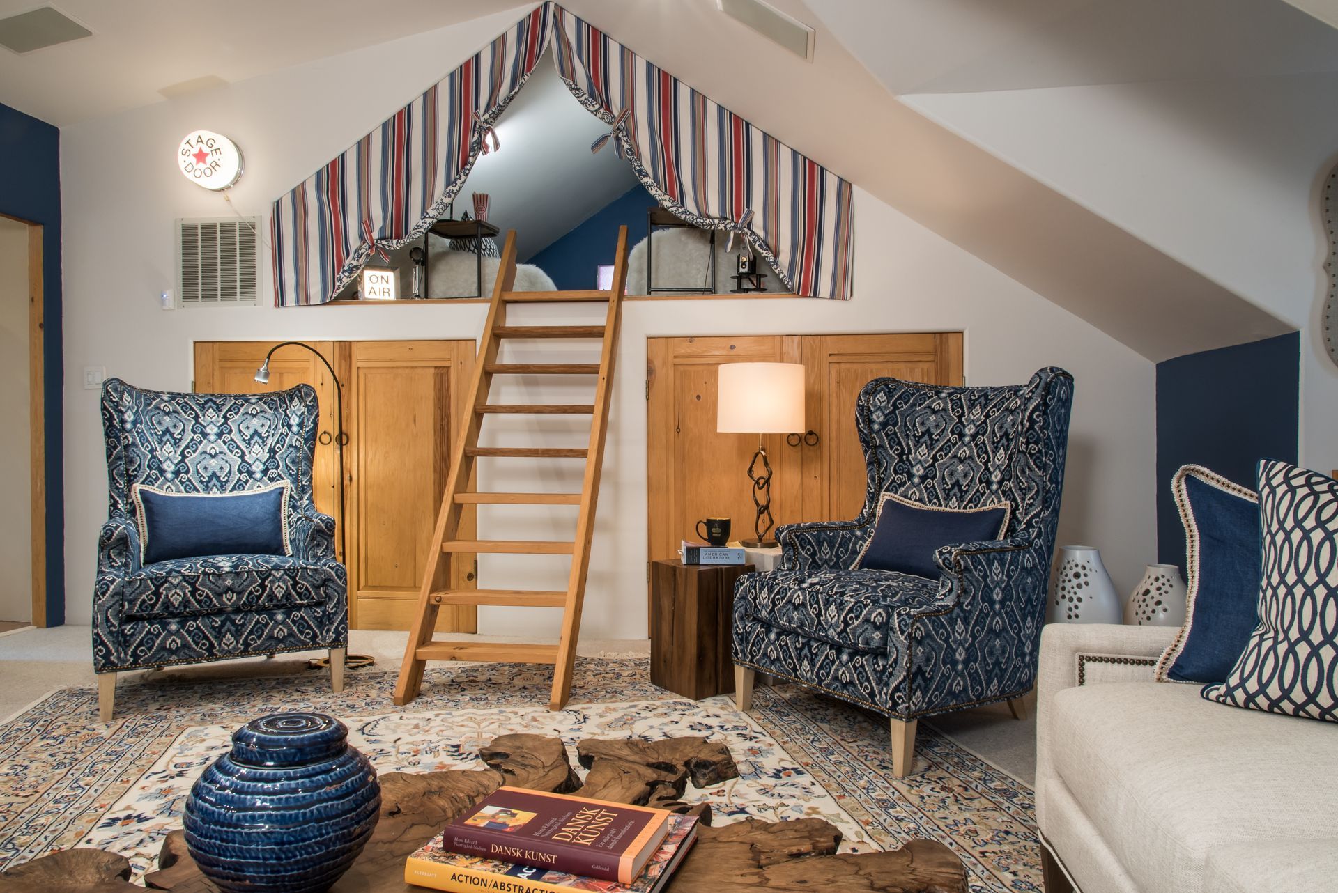 Cozy loft living room with blue chairs, ladder to mezzanine, patterned rug, and warm lamp lighting