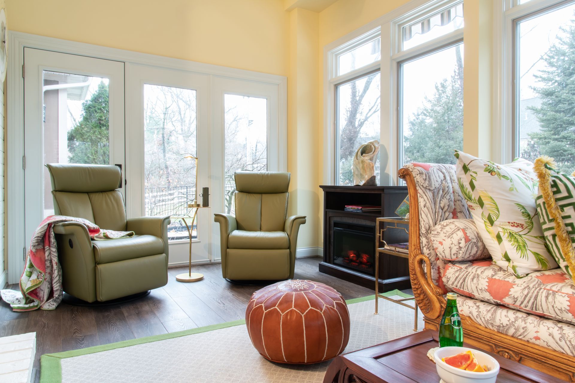 Bright living room with large windows, two chairs, sofa, fireplace, and an ottoman near the center.