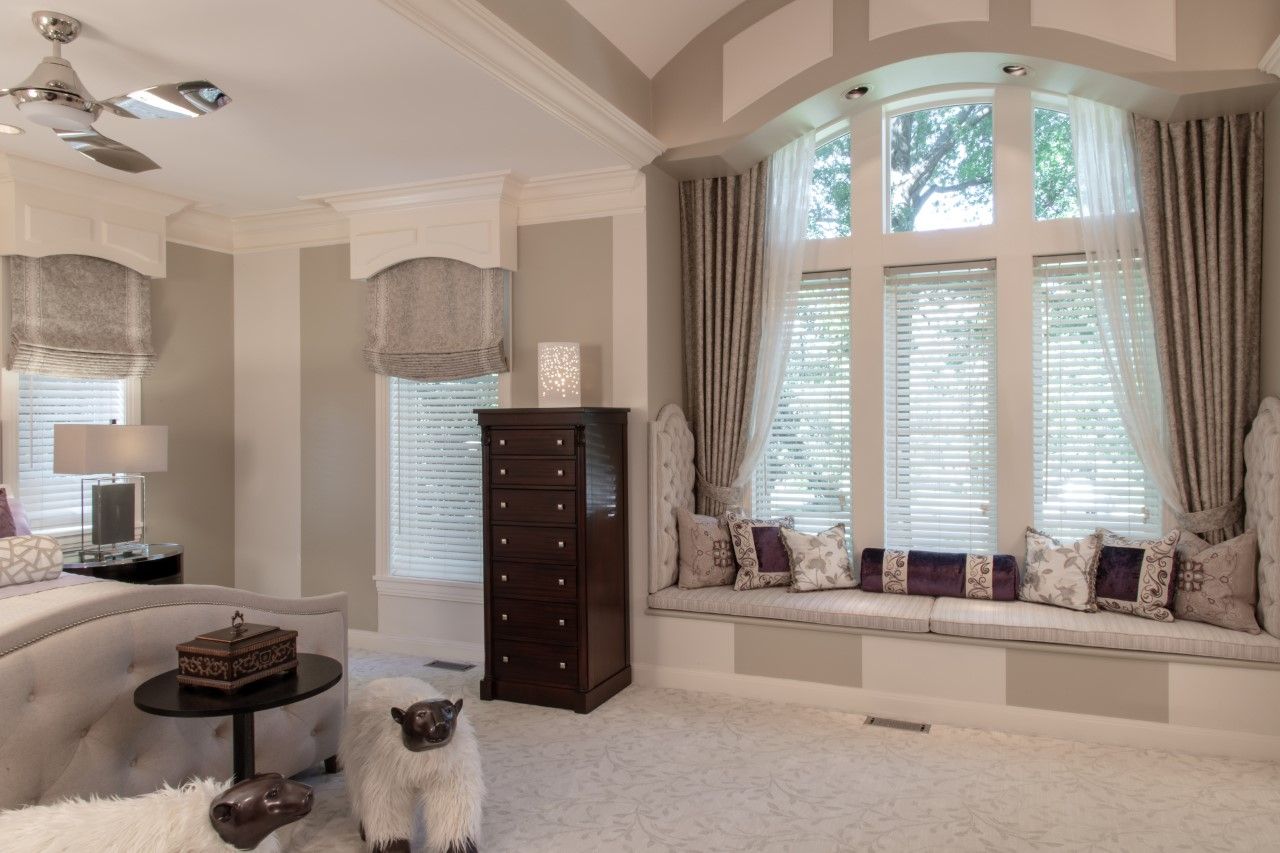 Bright beige bedroom with large bay window seat, dresser, chandelier, and round ottoman.
