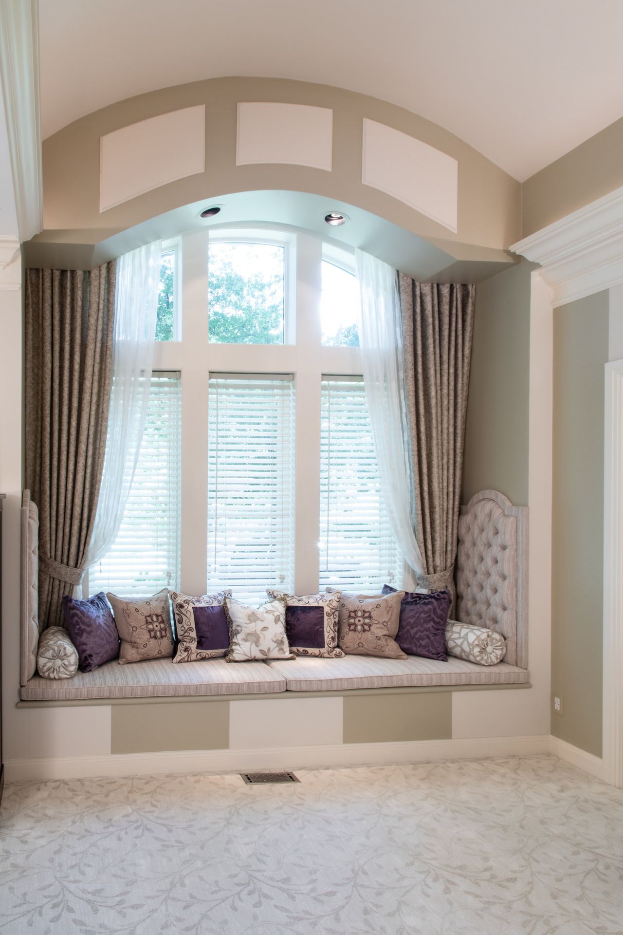 Built-in window seat beneath arched window, with taupe curtains and patterned cushions in a bright room