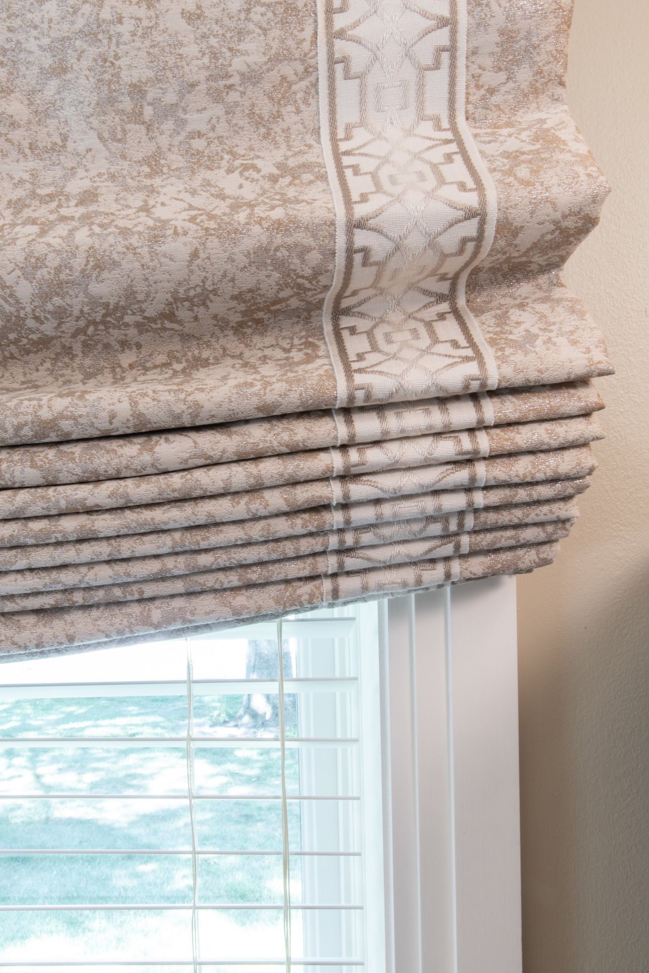 Beige patterned Roman shade partially raised over a window, showing the white frame and view outside