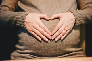 Hands forming a heart shape over a pregnant belly in a brown sweater.