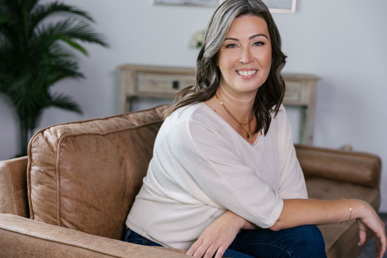 Woman sitting on a tan couch, smiling in a bright living room with a plant and lamp in the background