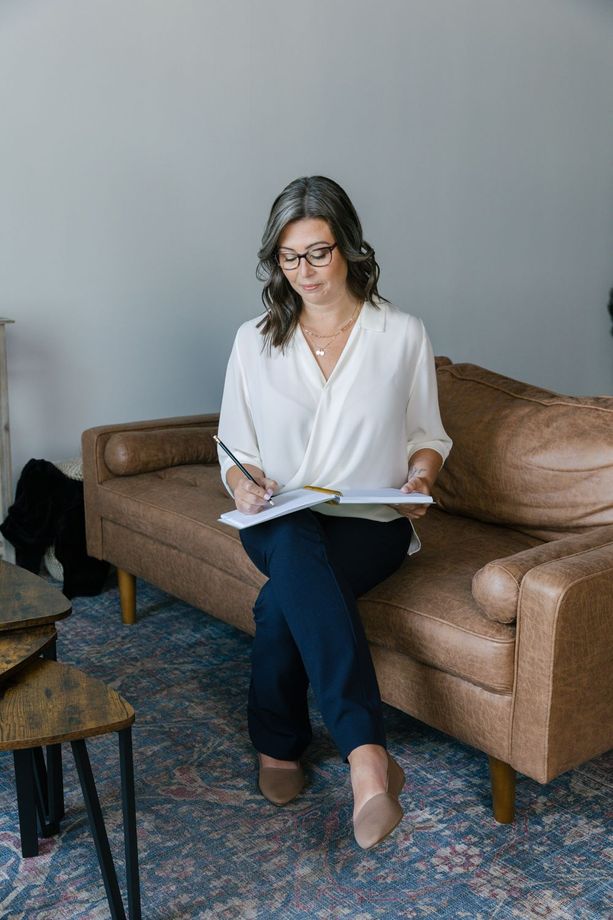 Woman seated on a brown sofa, reading and writing in a notebook beside a patterned rug.