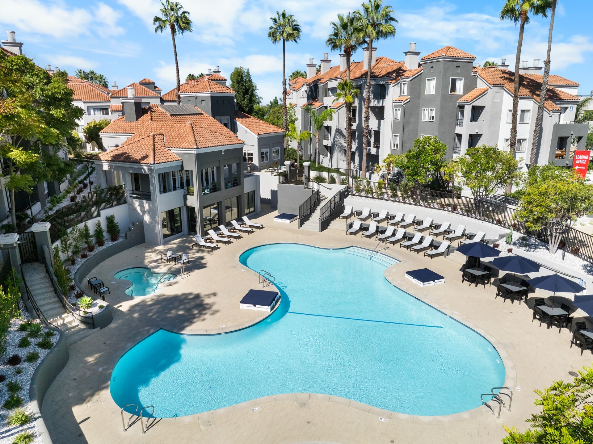 Pool area at apartment complex with lounge chairs, tables and umbrellas. Buildings in the background.