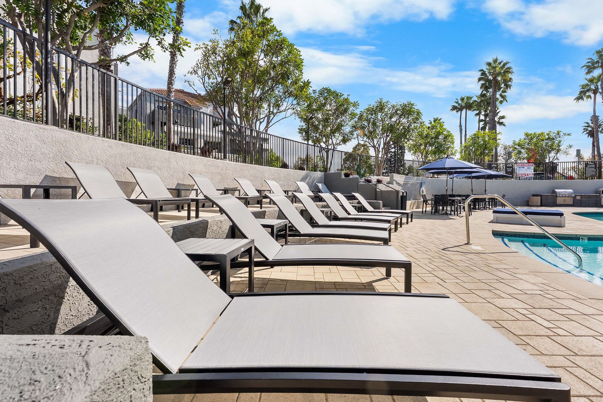 Lounge chairs by a pool on a sunny day. Trees and buildings in background.