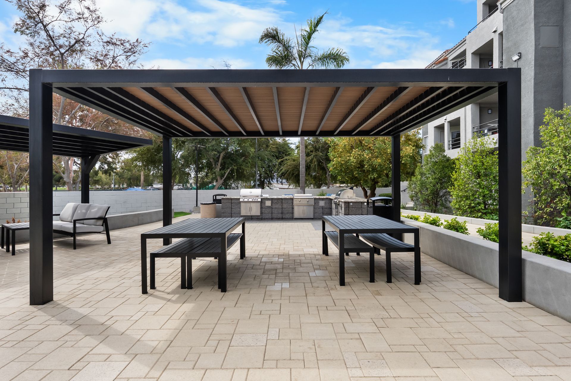 Patio with black pergola, tables, benches, and outdoor kitchen. Beige stone flooring with trees and building in the background.