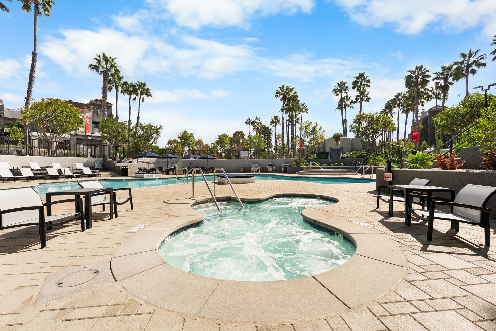 A hot tub and pool area, surrounded by palm trees, and lounge chairs, under a sunny sky.