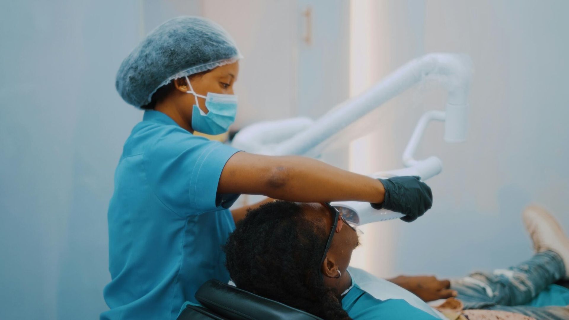 Dentist in blue scrubs and mask treating a patient in a dental chair under bright clinical lighting