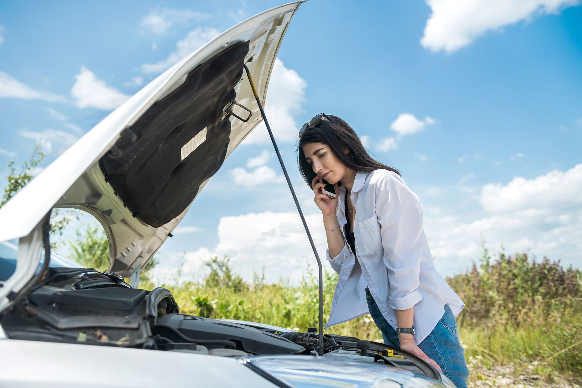 Woman checking under the hood of a broken-down car on a roadside under a cloudy sky