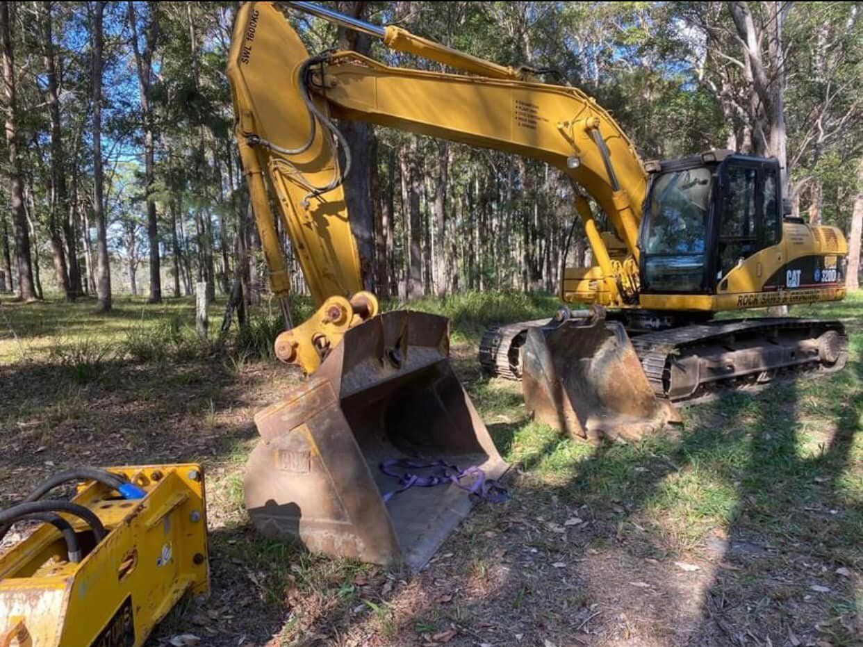 Yellow excavator with two buckets on grass in a wooded area — Ovannah Hire In West Kempsey, NSW