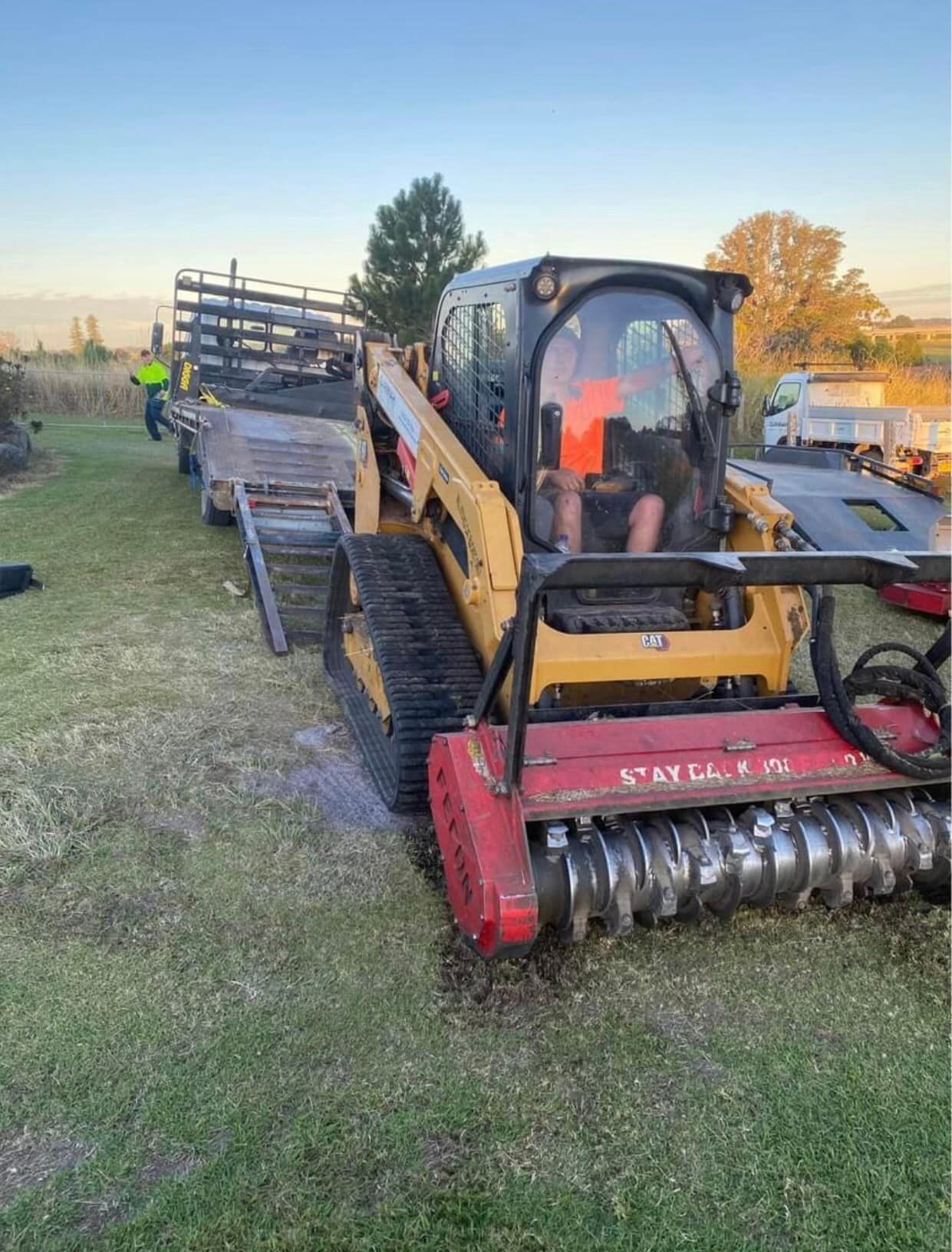 Yellow Skid Steer With Red Brush Cutter, Loading Onto Trailer in Grassy Area — Ovannah Hire In West Kempsey, NSW