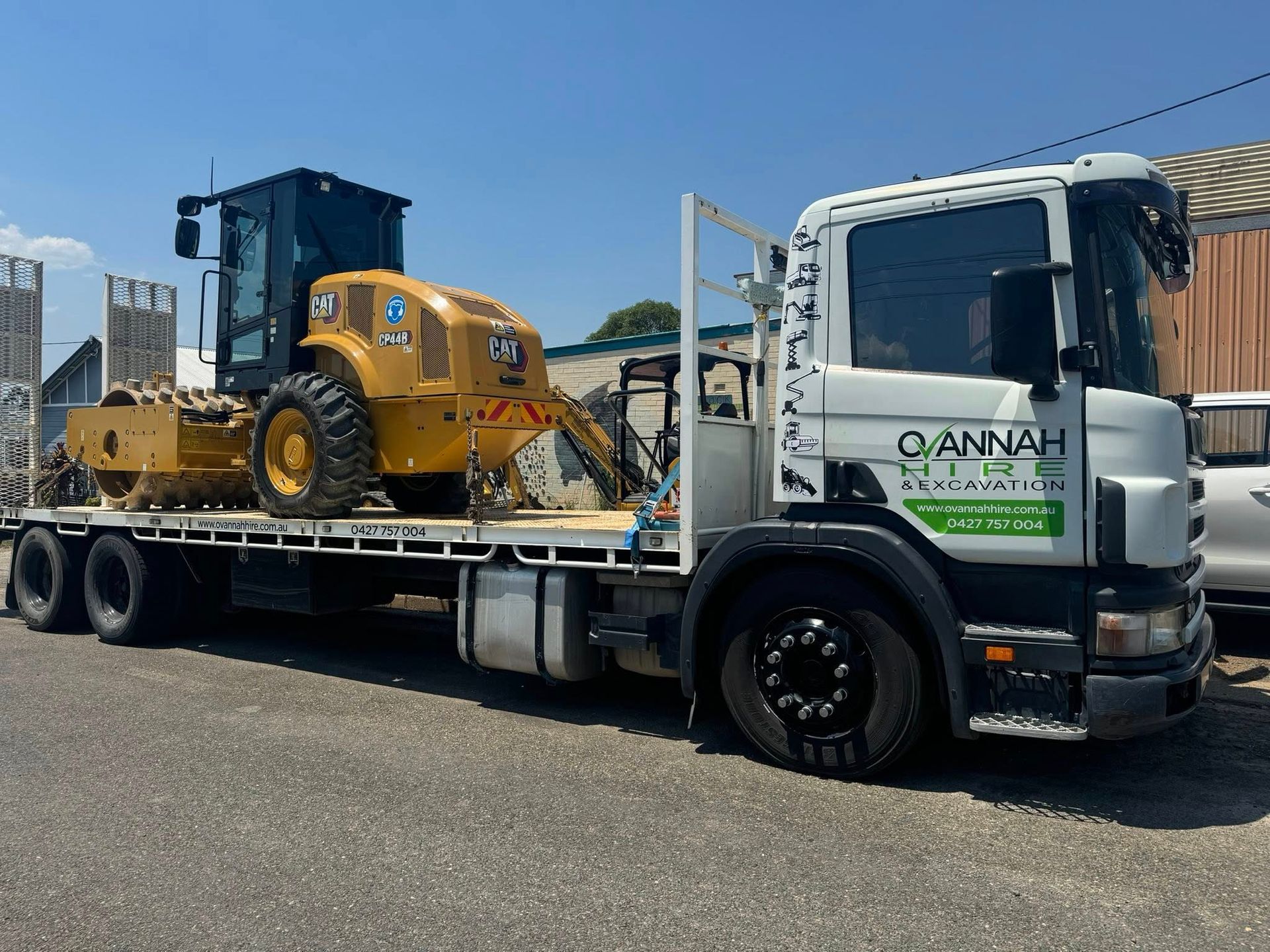 Flatbed Truck With a Yellow Cat Compactor on Its Bed — Ovannah Hire In West Kempsey, NSW