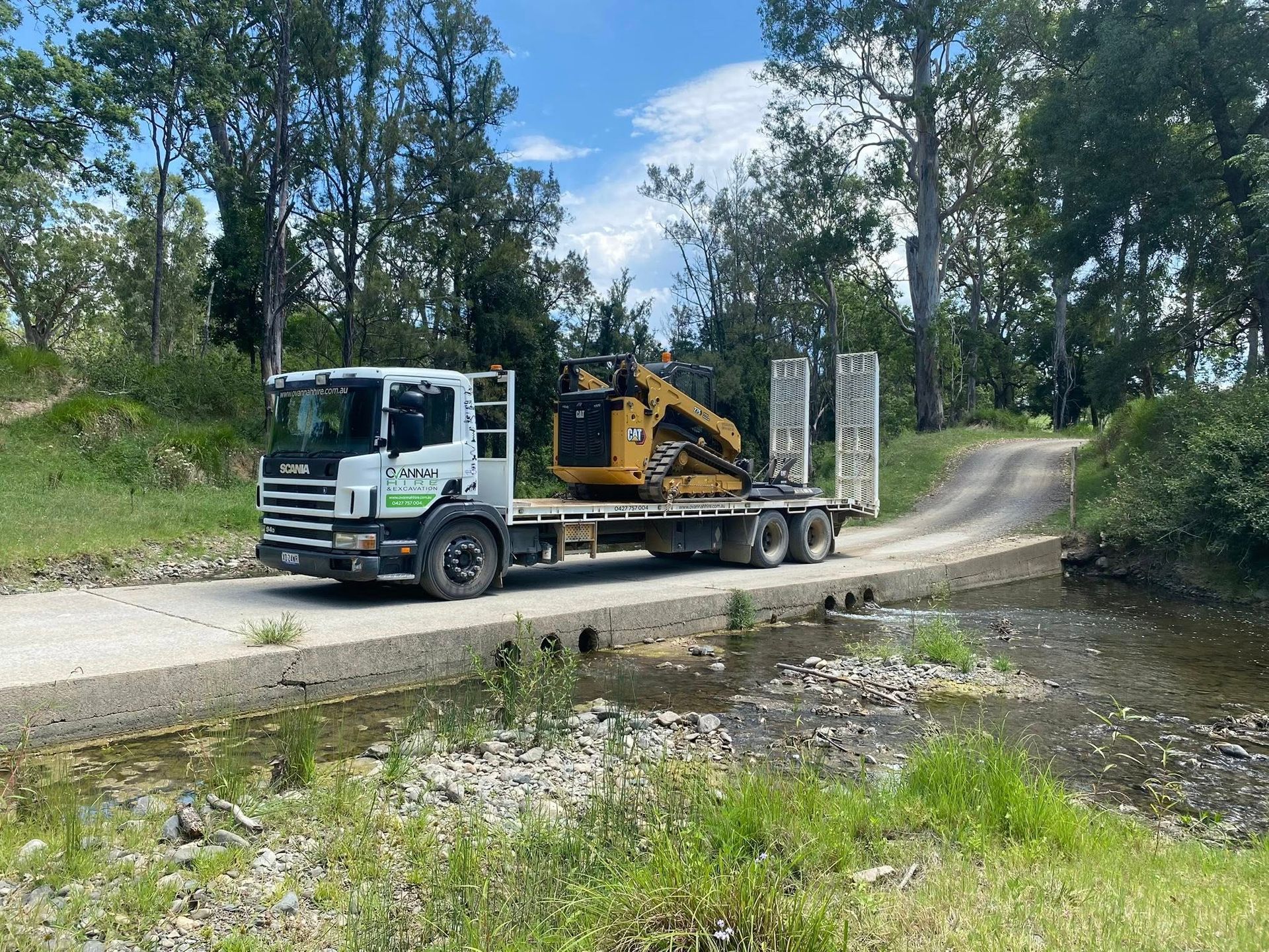 Truck Carrying an Excavator Crosses a Shallow Creek on a Concrete Bridge — Ovannah Hire In West Kempsey, NSW