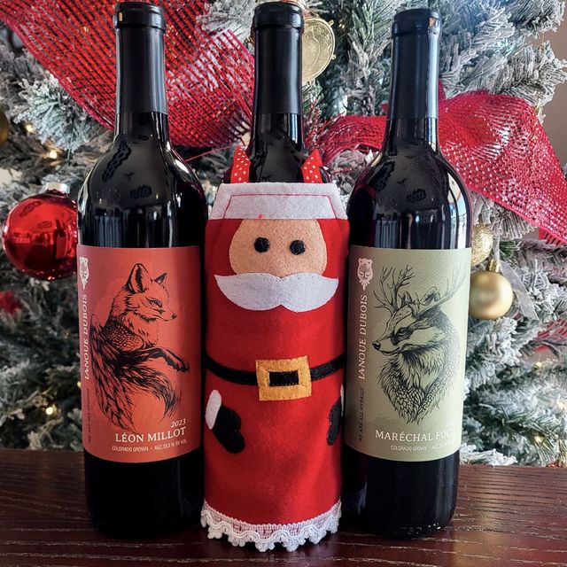 Three wine bottles with festive labels beside a Santa decoration and holiday ornaments on a frosted tree