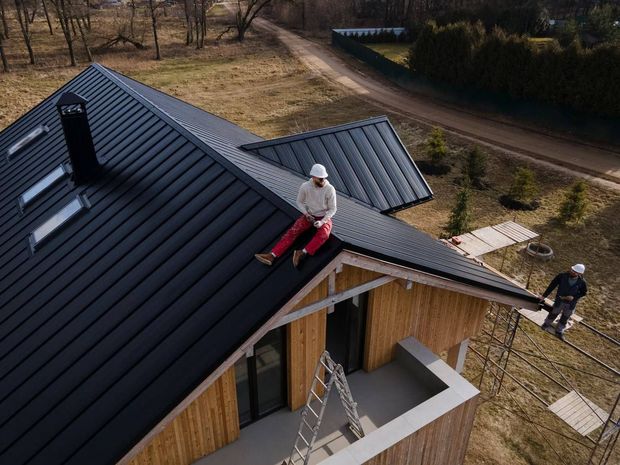 Two workers in safety helmets installing a dark metal roof on a new house with wooden siding in a rural setting.