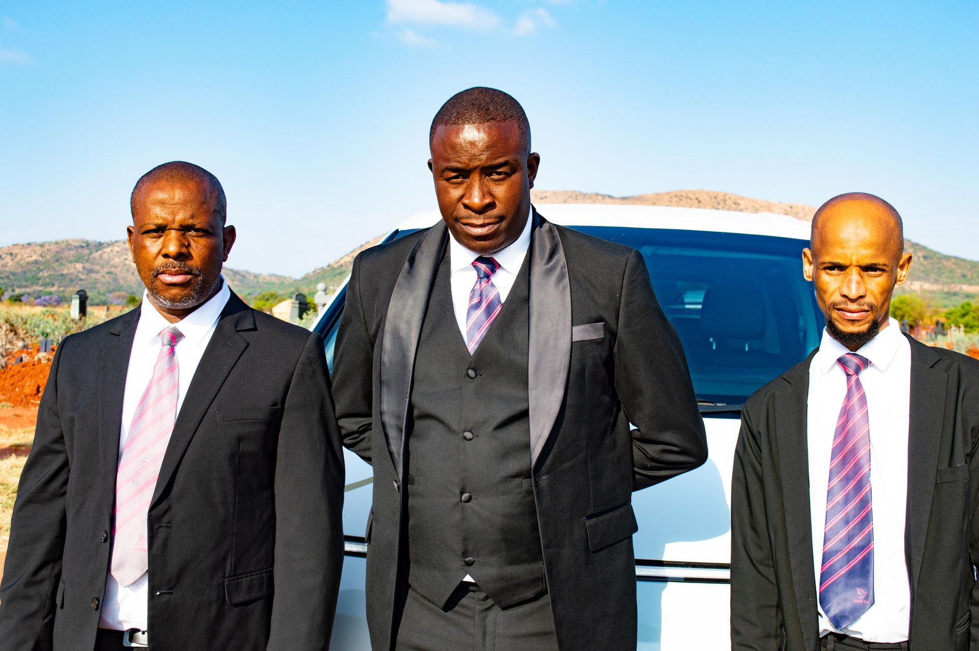 Three men in suits stand in front of a white vehicle against a sunny backdrop.